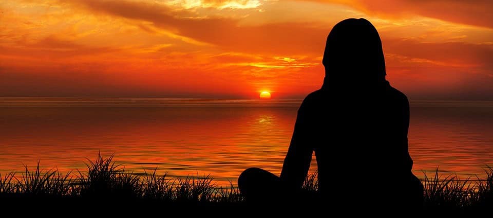 Sunset over calm ocean with silhouette of a girl sitting on grass, peaceful and calming scene.